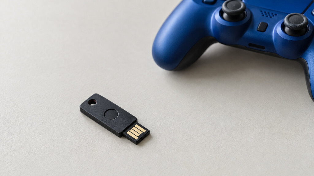 Crni USB hardverski sigurnosni ključ na sivoj podlozi ispred plavog PS4 kontrolera